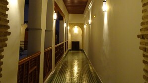 Um corredor com piso xadrez, portas em arco e vigas de madeira no teto.