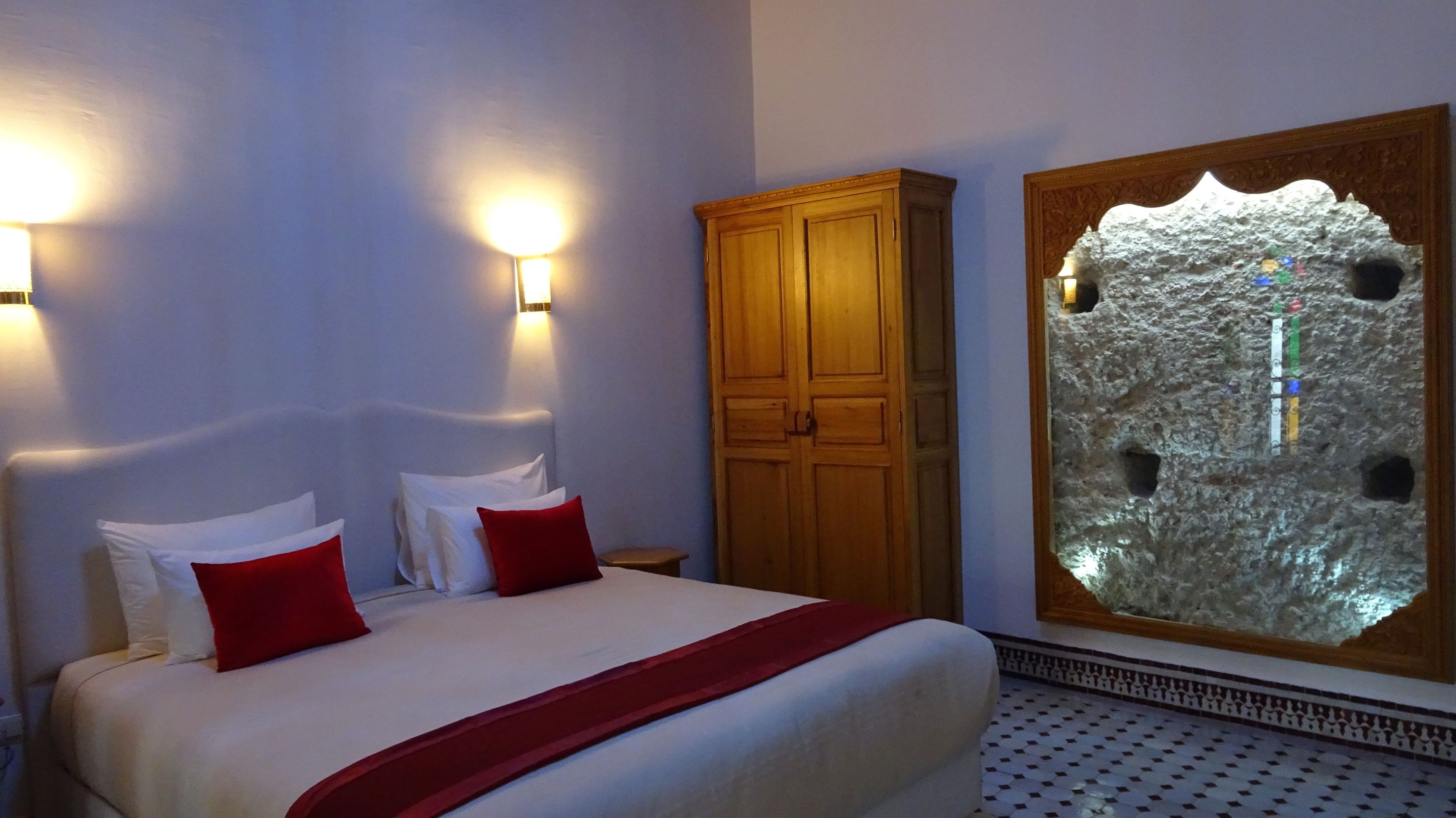 A hotel room with a bed, two wall-mounted lights, a wooden wardrobe, and a decorative stone wall.