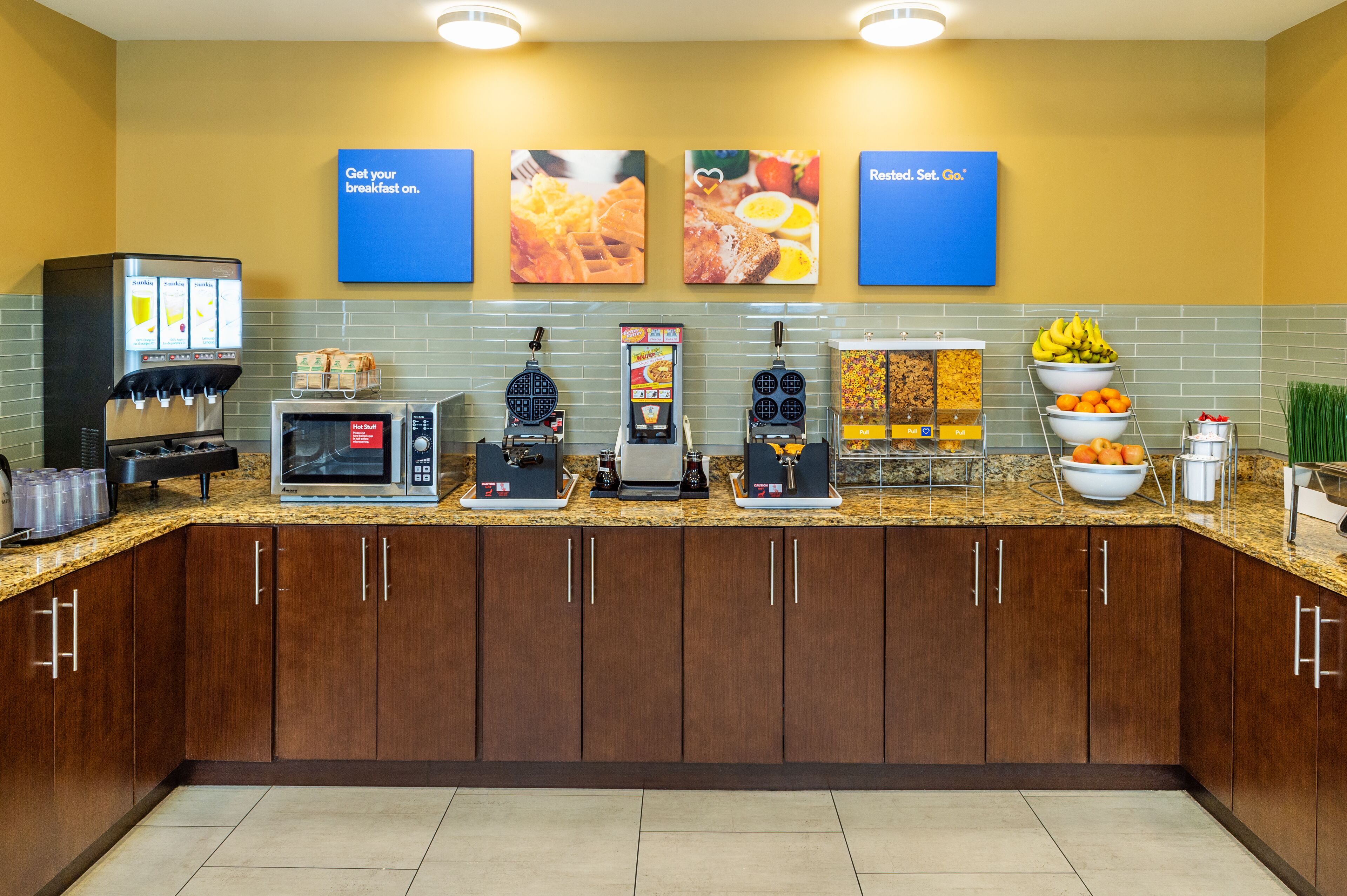 A breakfast bar with various food and drink options, including a waffle maker, coffee machine, and a display of fresh fruits and vegetables.