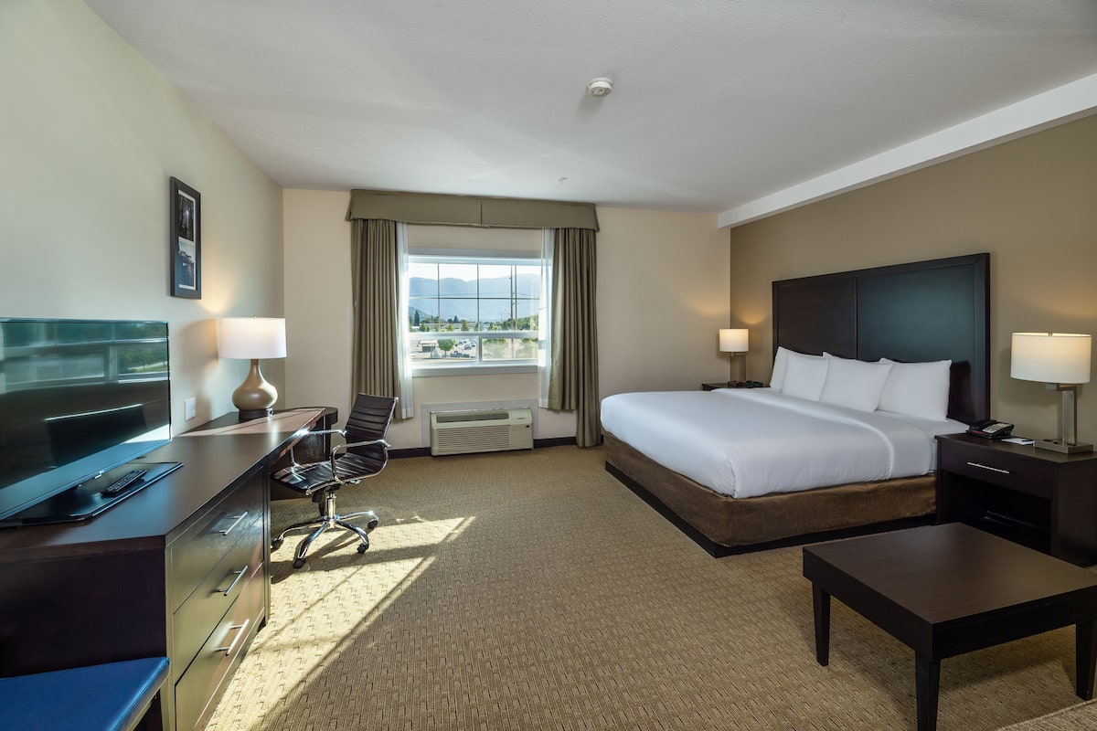 A hotel room with a large bed, a desk with a TV, a chair, a small table, and a window with curtains.
