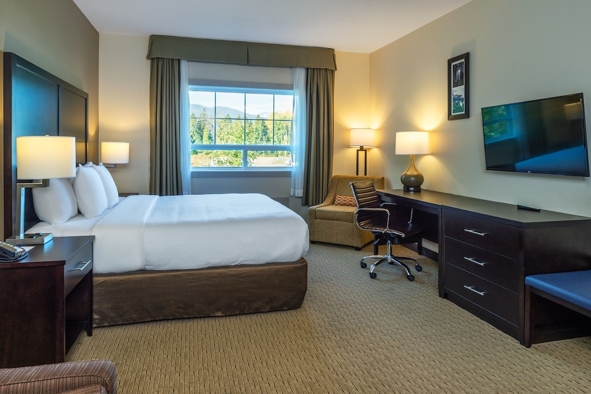 A hotel room with a large bed, two bedside tables, a desk with a chair, a TV, and a window with curtains.