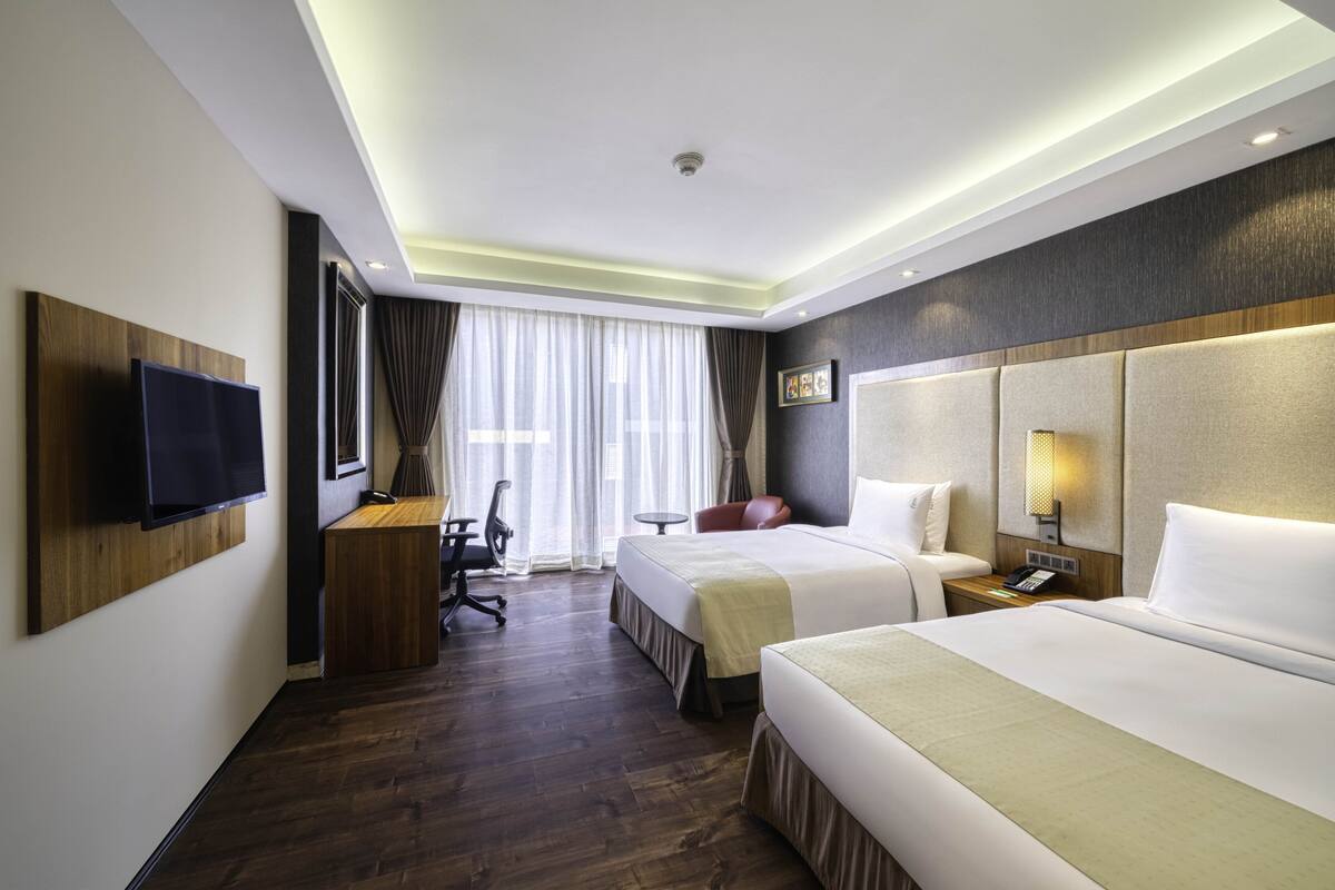 A hotel room with two beds, a desk, a chair, a TV, and a window with curtains.