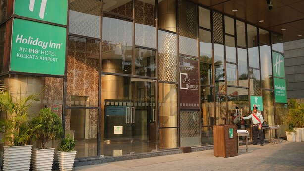 Exterior del Holiday Inn Kolkata Airport con amplios ventanales de vidrio, un guardia de seguridad y un cartel que indica que es un hotel IHG.