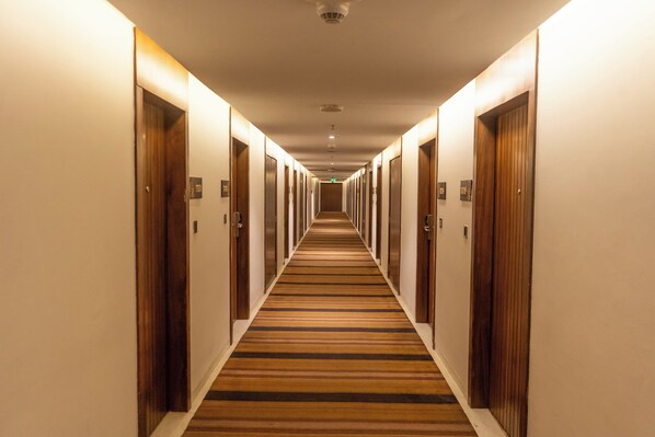 Un long couloir étroit, typique d’un hôtel, avec des portes en bois de chaque côté et un tapis à rayures.