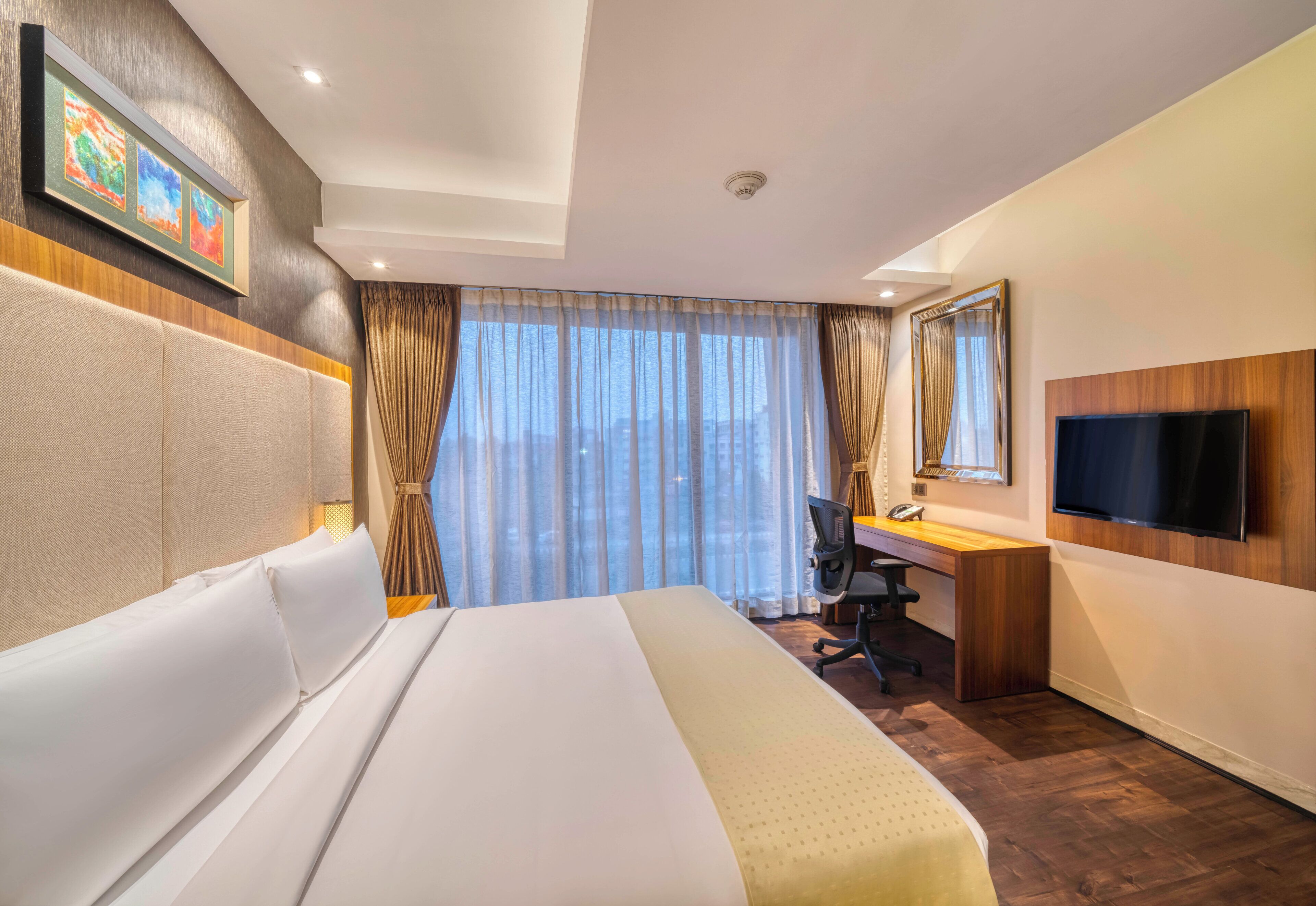 Ein Hotelzimmer mit Bett, Schreibtisch, Stuhl, Fernseher und einem großen Fenster mit Vorhängen.