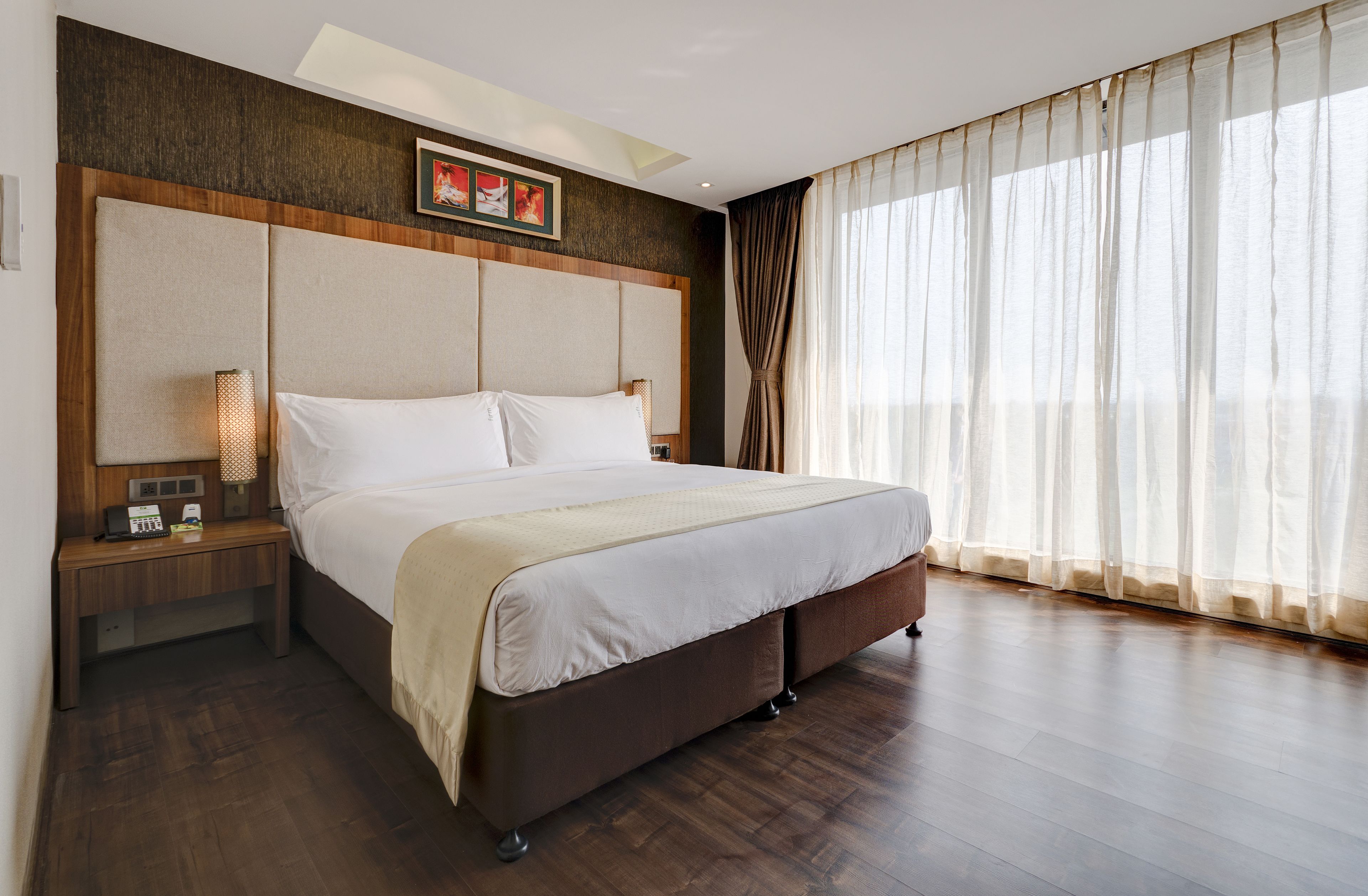 A hotel room with a large bed, wooden headboard, a nightstand with a lamp, and sheer curtains on the window.