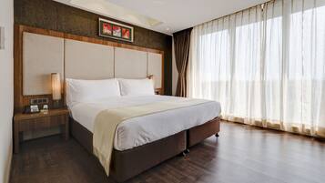 A hotel room with a large bed, wooden headboard, a nightstand with a lamp, and sheer curtains on the window.