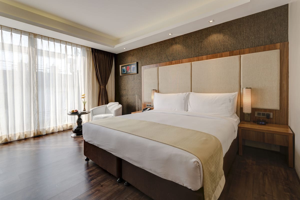 A hotel room with a large bed, a bedside table with a lamp, a chair, and a view of the outdoors through sheer curtains.