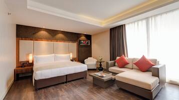A hotel room with a large bed, a sofa, a coffee table, and a chair. There is a window with curtains, a wall-mounted clock, and a small table with a vase of flowers.