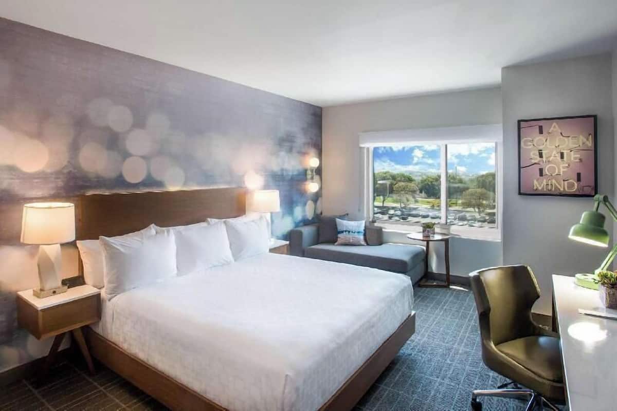A hotel room with a large bed, a desk, a chair, a small table, and a view of the outdoors.
