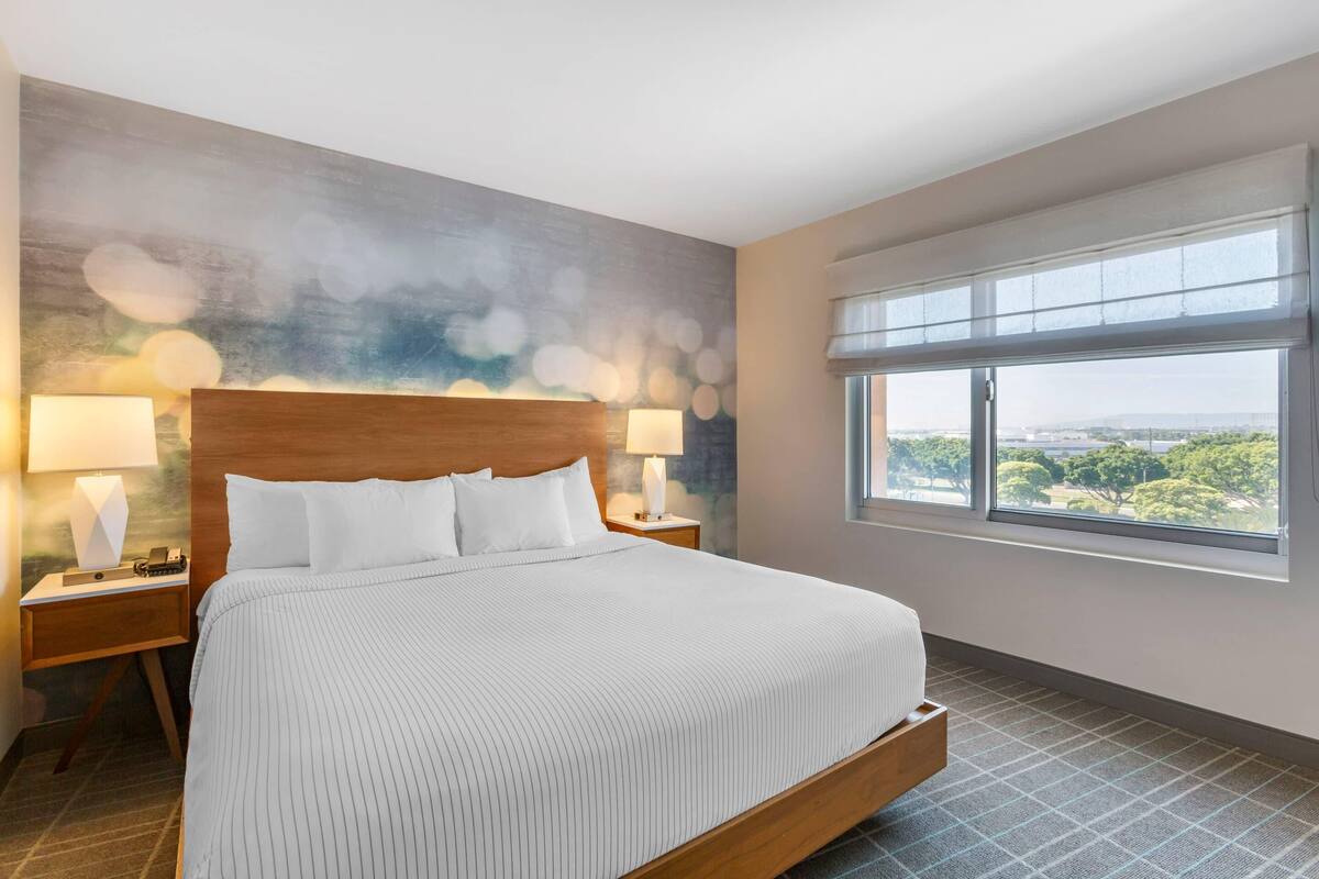 A hotel room with a large bed, two bedside tables with lamps, a view of greenery through the window, and a textured wall mural.