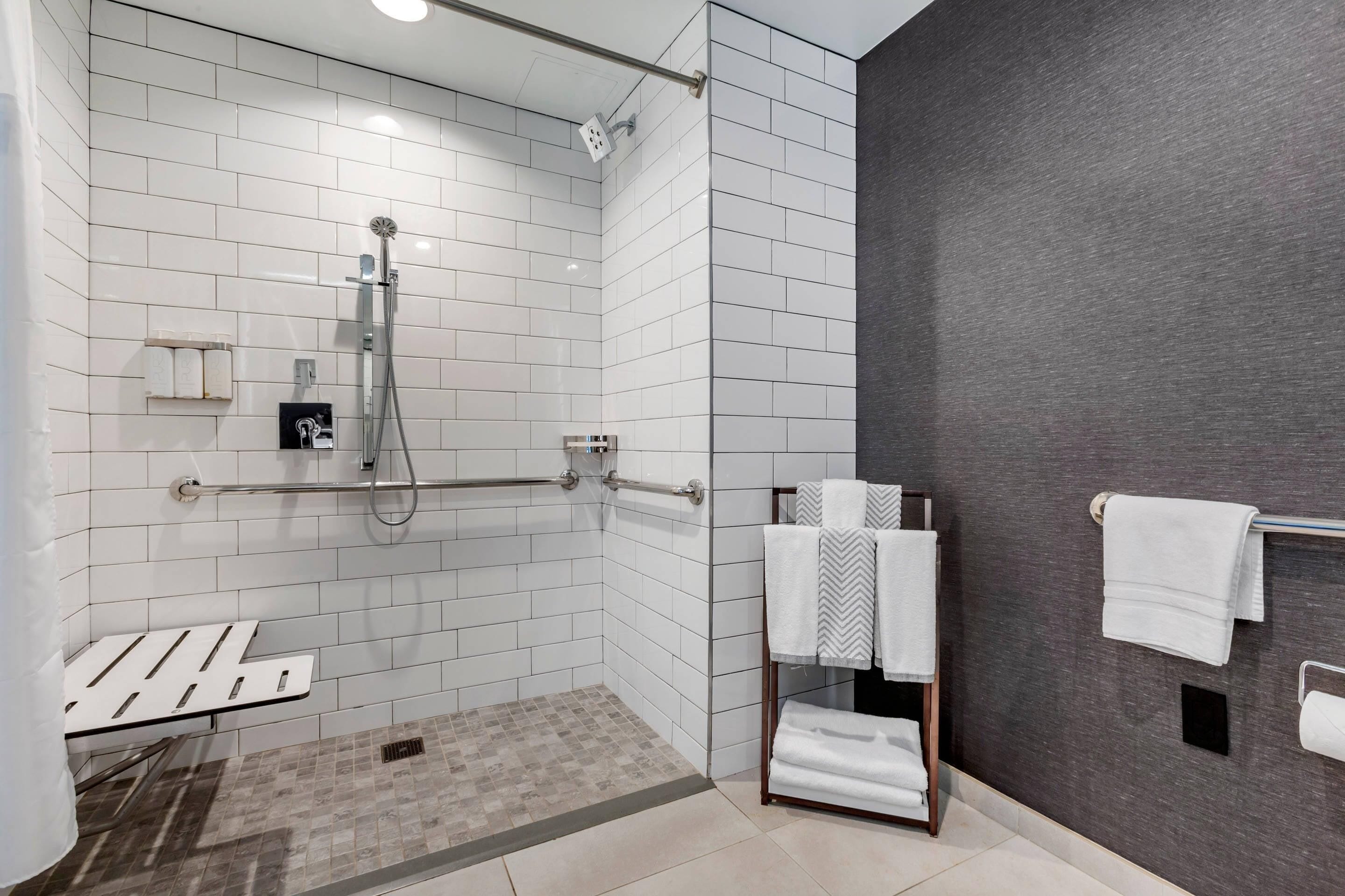 A modern bathroom with white tiled walls, a shower area with a glass door, a towel rack, and a small shelf with toiletries.
