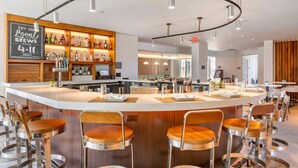 A modern bar area with a central island, bar stools, and a variety of liquor bottles. - Cambria Hotel LAX (El Segundo)