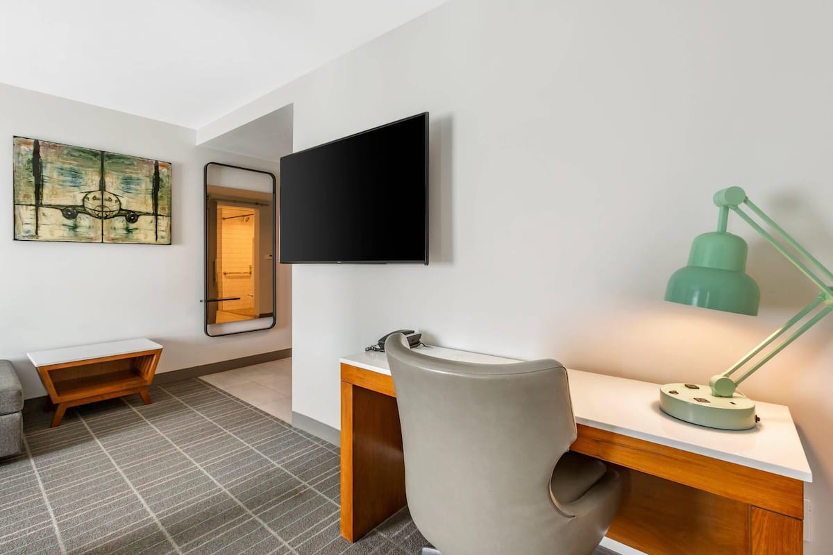 A modern hotel room with a flat-screen TV mounted on the wall, a desk with a chair, a small wooden side table, and a green desk lamp.
