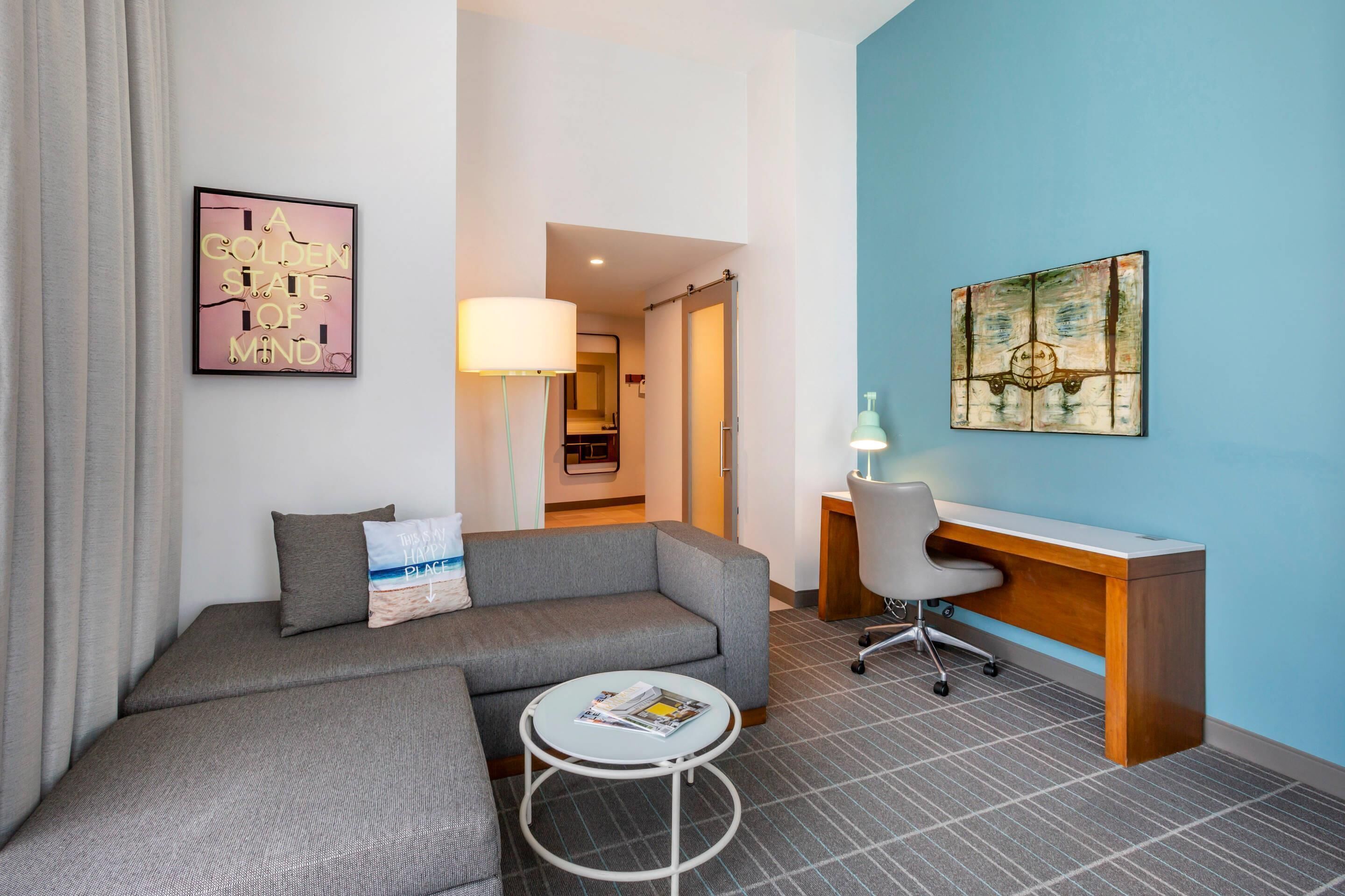Una habitación de hotel moderna con un sofá gris, una pequeña mesa redonda, un escritorio de madera y una pared azul decorativa.