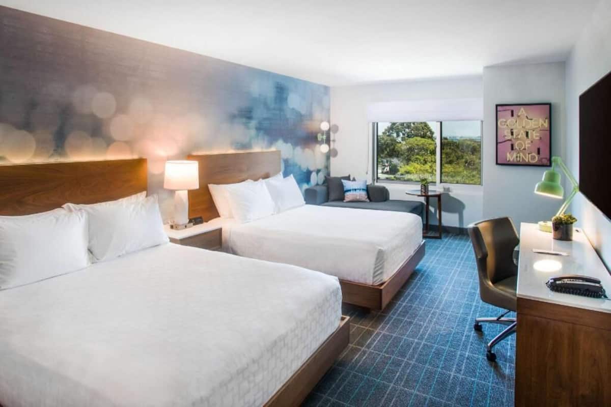 A hotel room with two beds, a desk, a chair, a lamp, and a large window with a view.