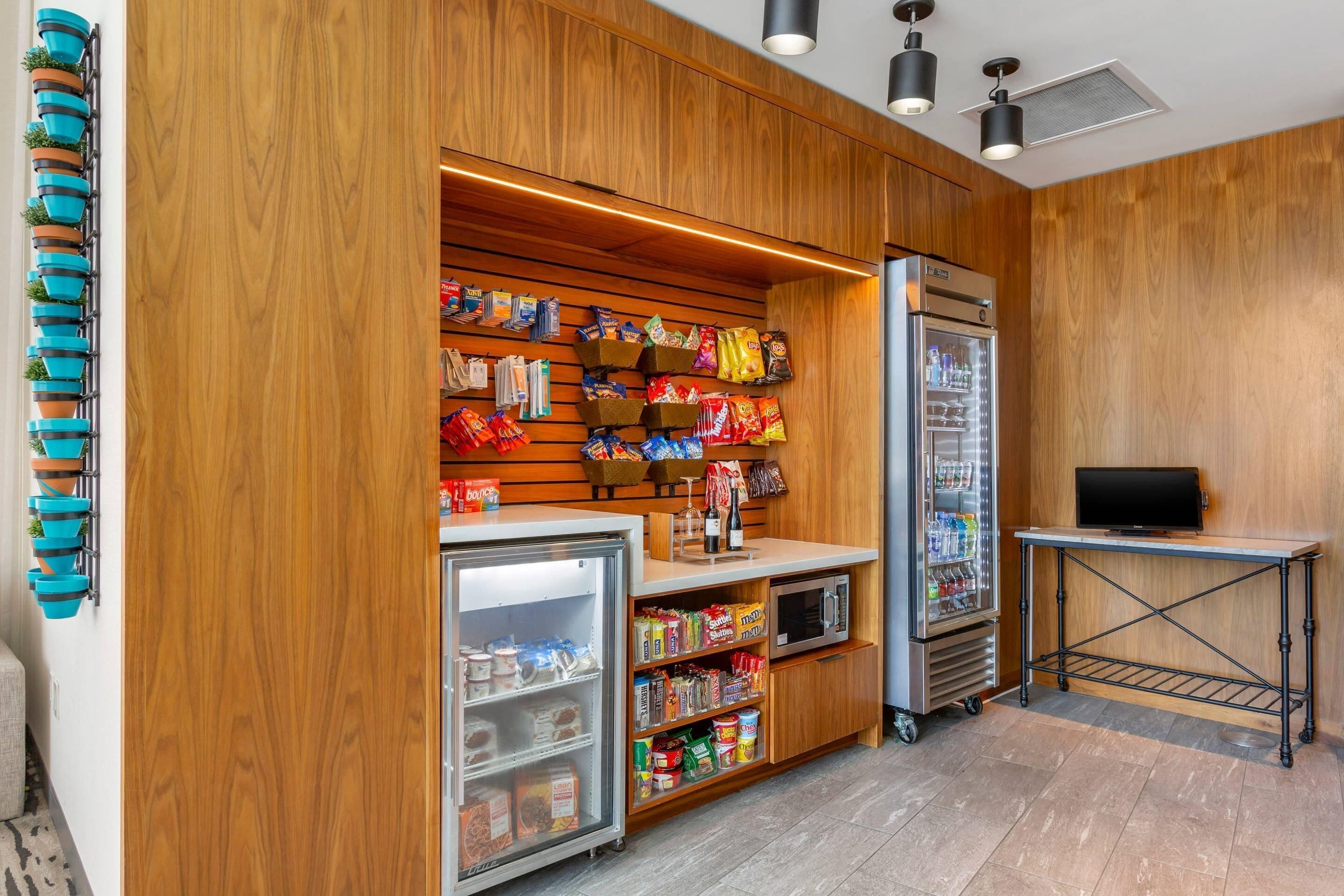 Una habitación pequeña con paneles de madera, un refrigerador, un microondas y una estantería llena de aperitivos y bebidas.