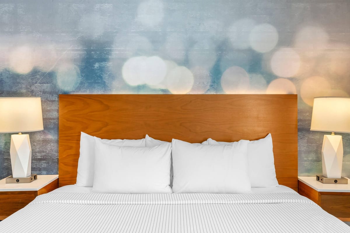 A neatly made bed with white linens and pillows, flanked by two bedside tables with lamps, against a backdrop of a textured wall with a bokeh effect.