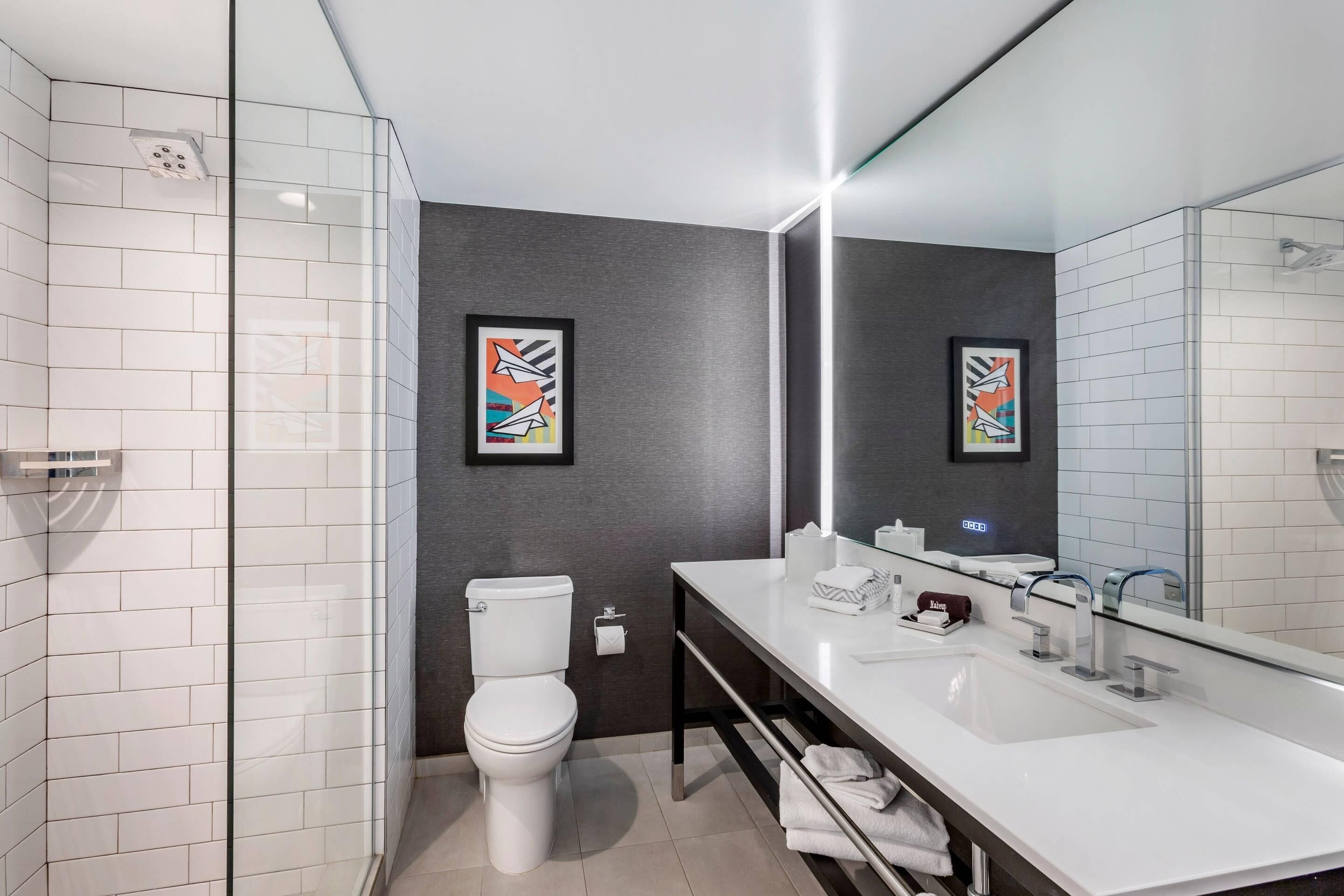 A modern bathroom with a white sink, a large mirror, and a toilet. There are two framed artworks on the wall.