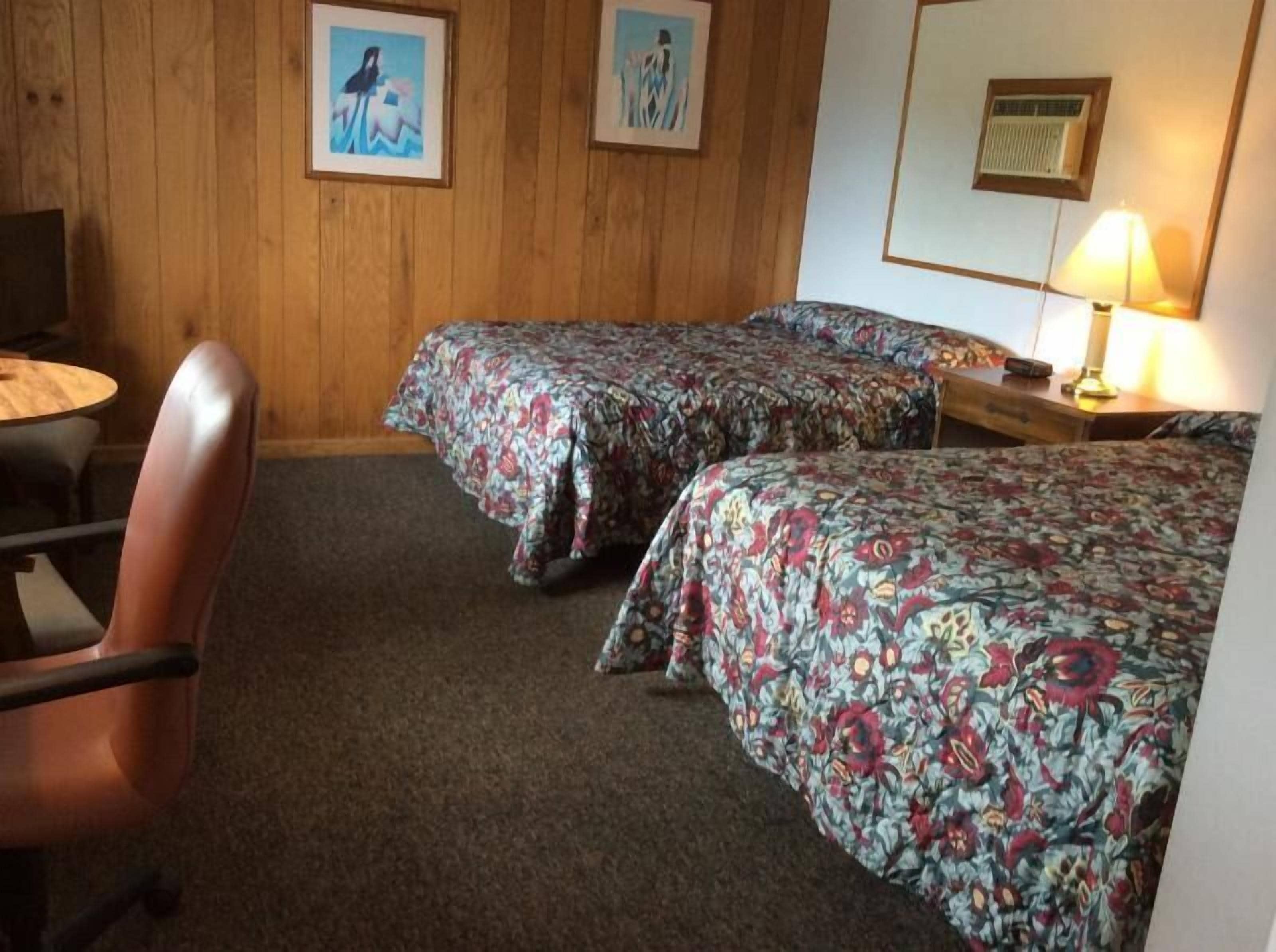 Ein Hotelzimmer mit zwei Betten, einem Holz-Kopfteil, einem Schreibtisch mit Lampe und zwei gerahmten Bildern an der Wand.
