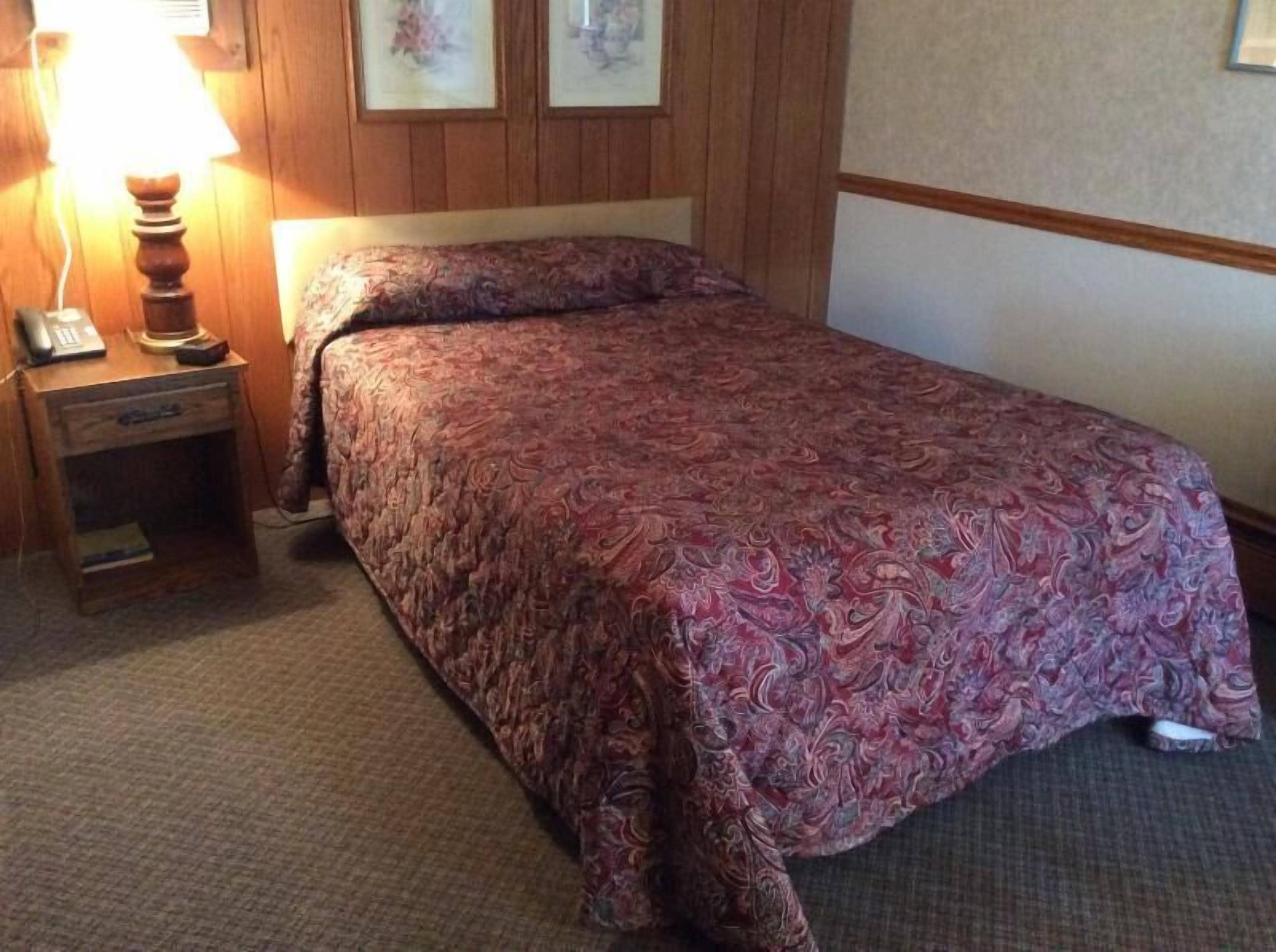 Een net opgemaakt bed met een bedsprei met patroon in een kamer met houten lambrisering en een klein nachtkastje met een lamp.