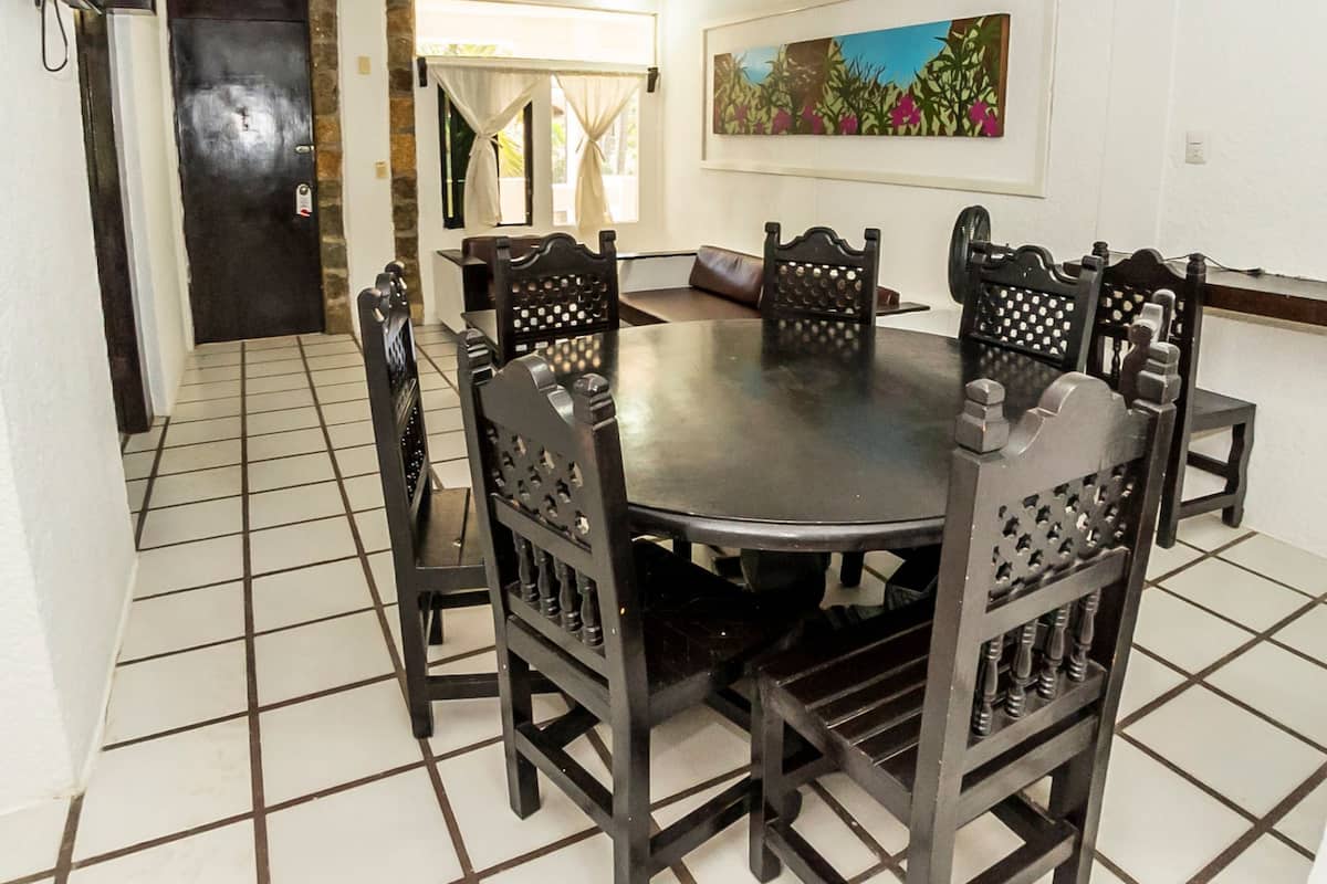 A dining area with a round table surrounded by six wooden chairs with intricate back designs. A painting hangs on the wall above the table.