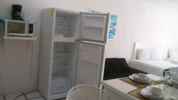 Una habitación pequeña con una cama blanca, un pequeño comedor con platos y vasos, un refrigerador blanco y un microondas en un estante.