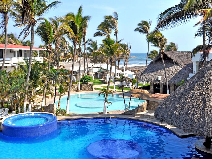 A resort with a large pool surrounded by palm trees and thatched-roof huts.