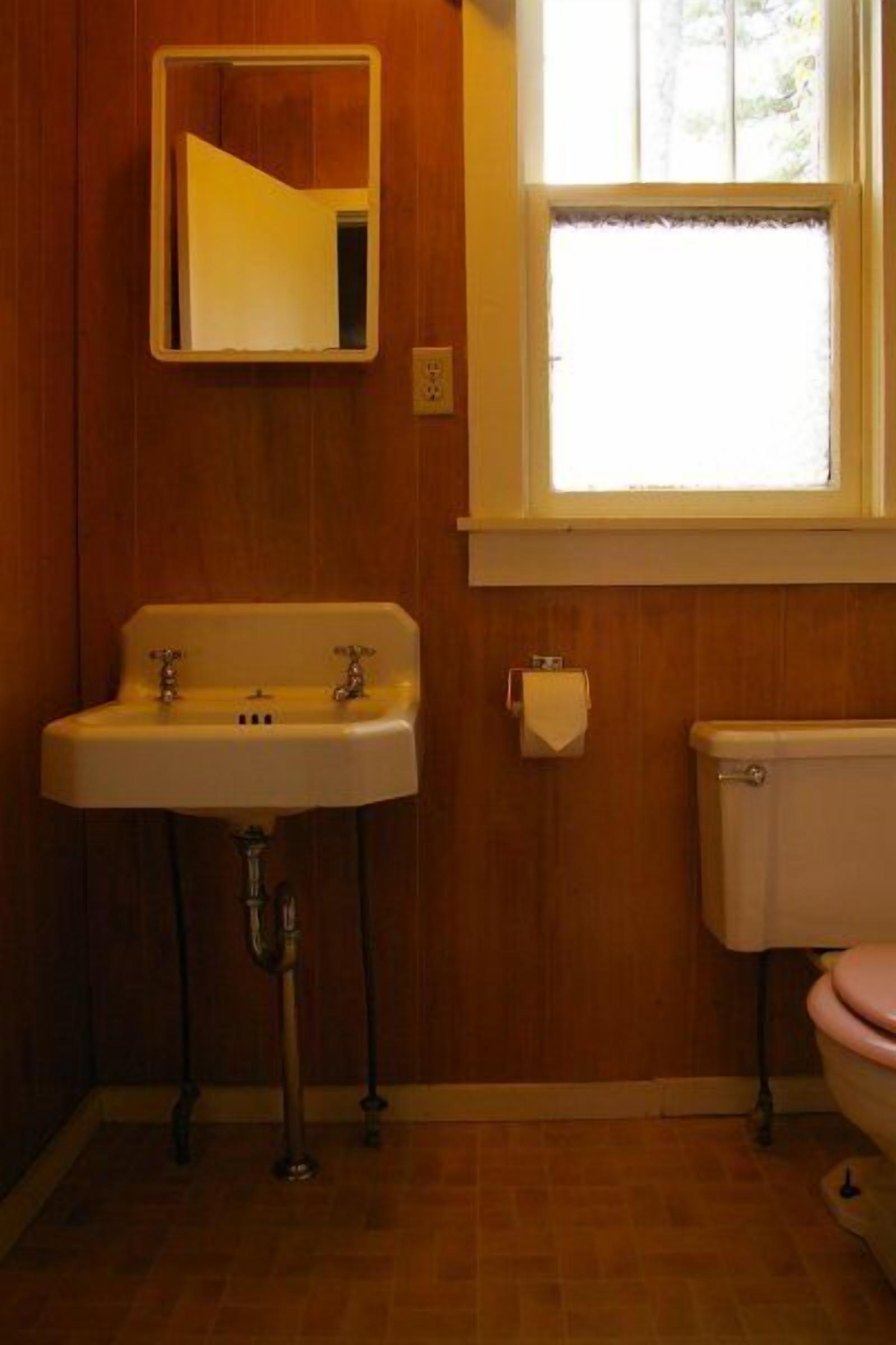 A bathroom with a wooden wall, a white sink with two faucets, a toilet, and a window above the sink.