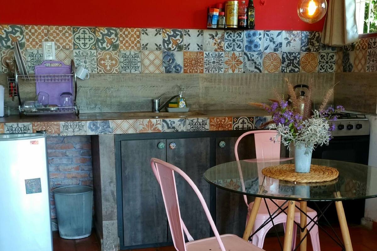 A kitchen with a red wall, tiled backsplash, and a round glass table with pink chairs.