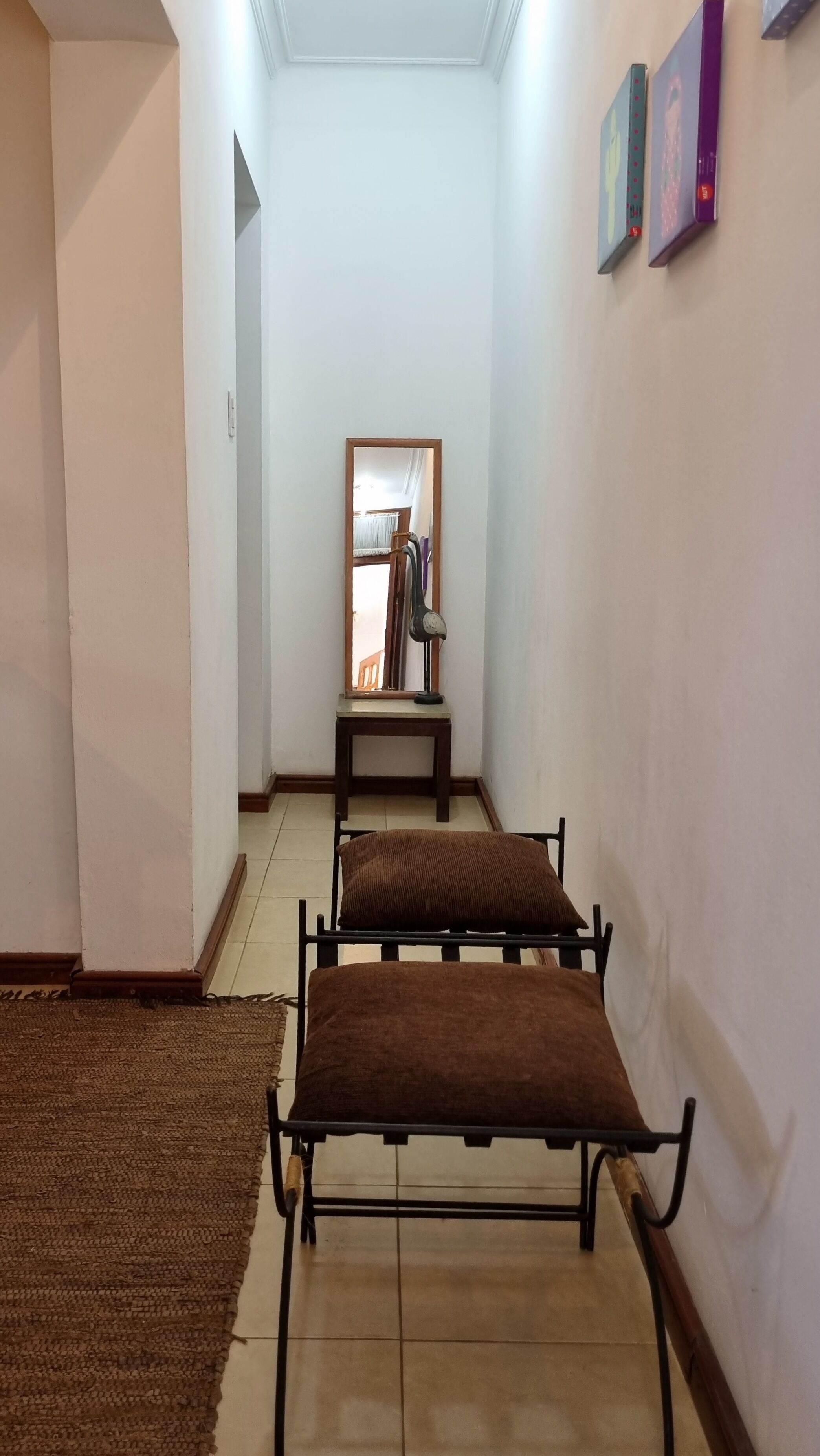 A narrow hallway with two black metal chairs with brown cushions, a wooden table with a mirror and books, and a brown rug on the floor.
