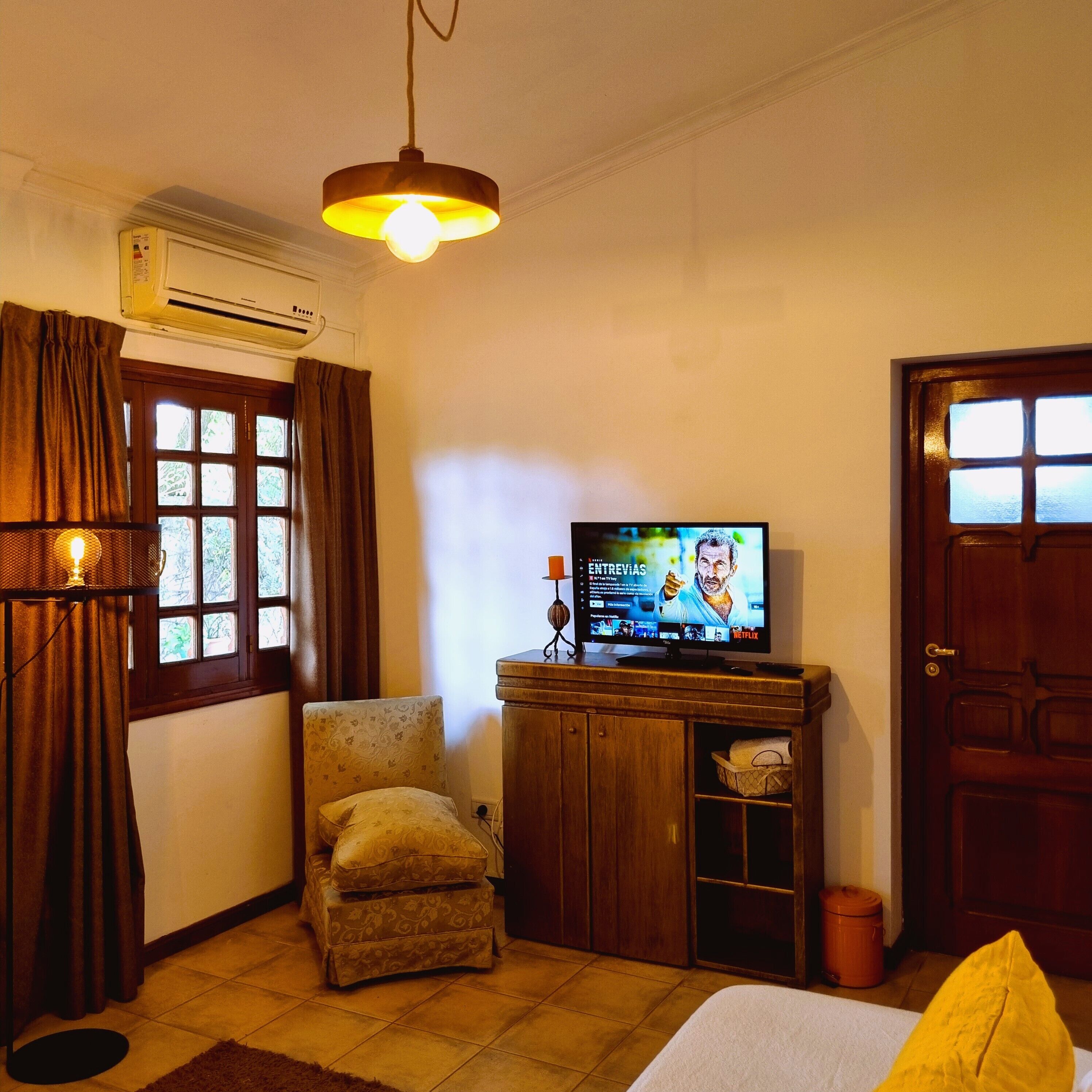 Una habitación con una cama, televisión, una silla, un armario de madera y una ventana con cortinas.