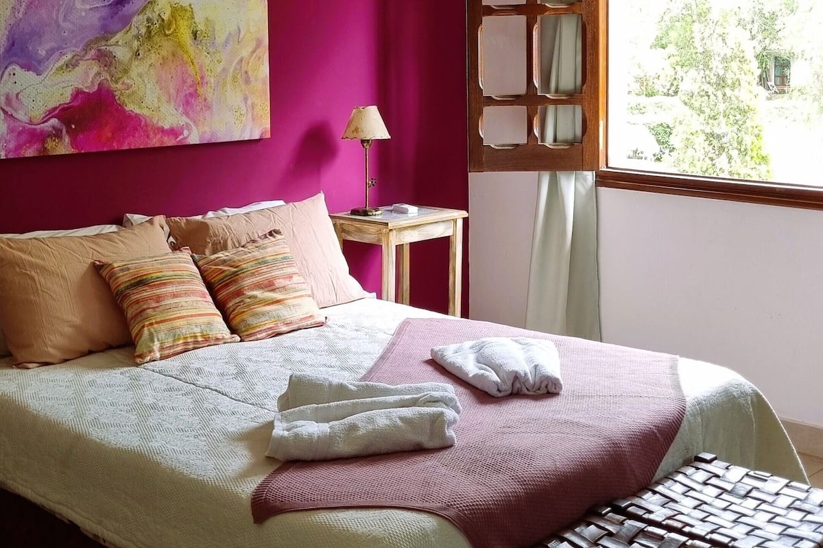 A bedroom with a large bed, a wooden bench, a nightstand, a lamp, and a window with a view of greenery.