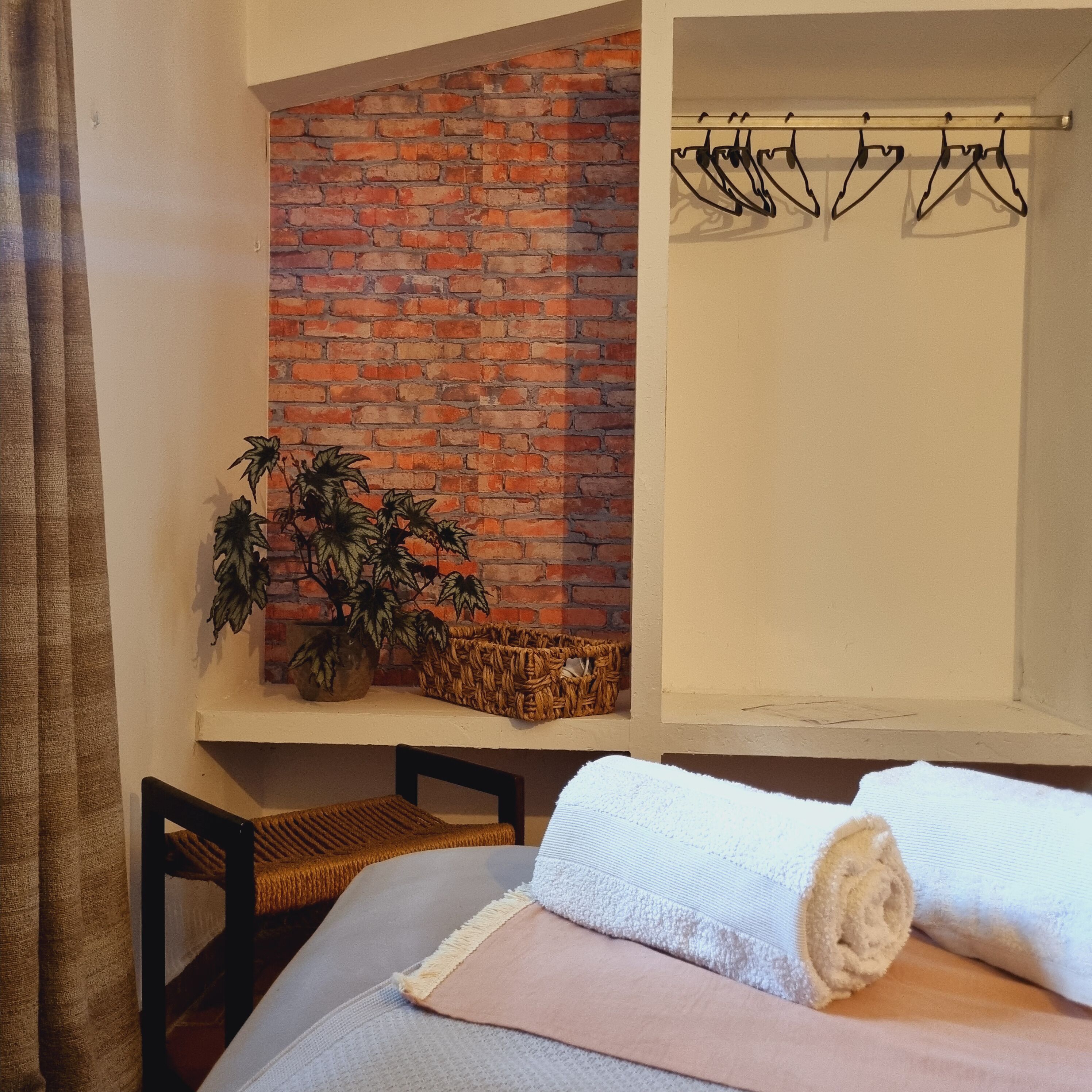 A bedroom with a brick wall, a plant, a hanging clothes rack, and a bed with a rolled-up towel.