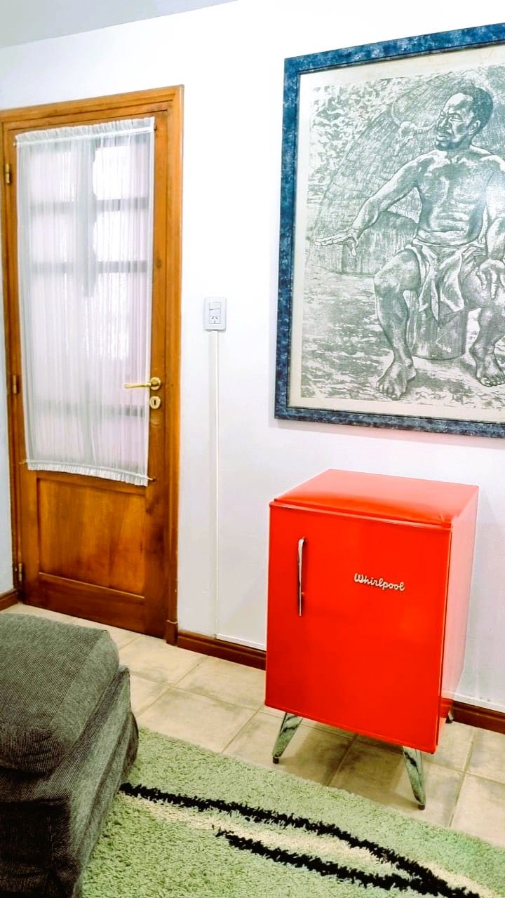 A room with a wooden door, a red Whirlpool refrigerator, a green sofa, and a framed artwork on the wall.