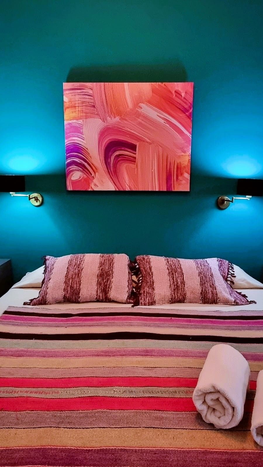 Hotellihuone, jossa on turkoosi seinä, suuri abstrakti maalaus, raidallinen vuodevaatteilla varustettu sänky ja kaksi yöpöytälamppua.