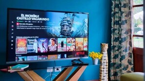 Ein Wohnzimmer mit einem großen Flachbildfernseher, der einen Film zeigt, einem Holztisch, zwei gelben Sesseln und einem Fenster mit gemusterten Vorhängen.