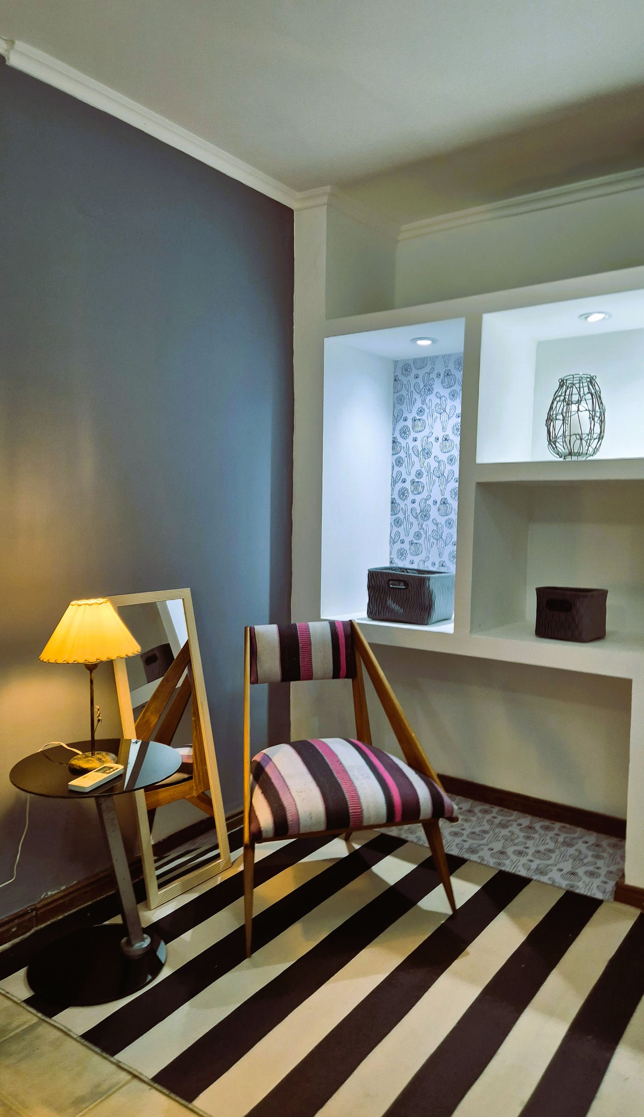 A modern living room with a striped rug, a wooden chair with a striped cushion, a lamp, and a shelf with decorative items.