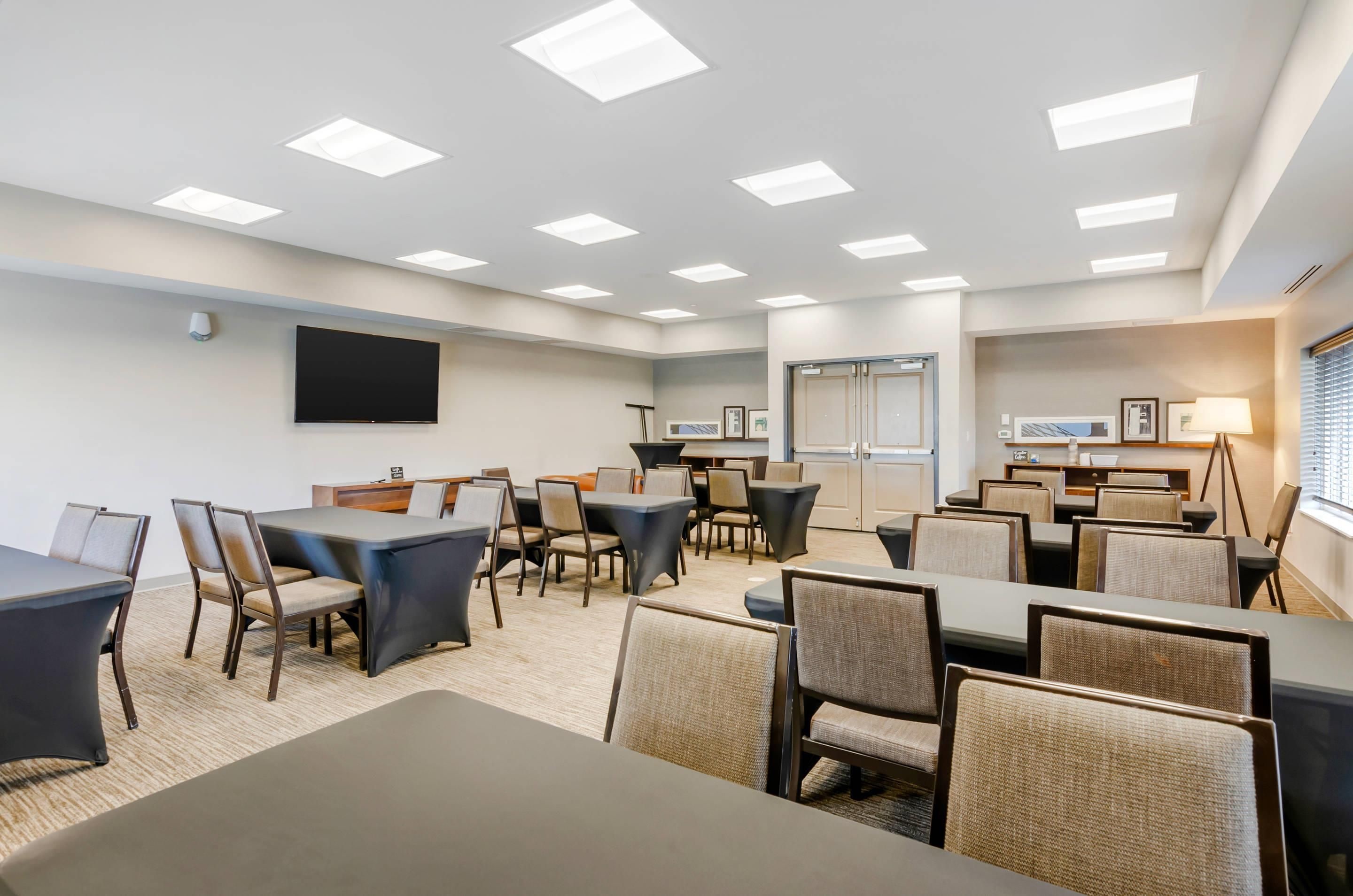 Une salle de conférence d’hôtel équipée de plusieurs tables et chaises, d’un téléviseur à écran plat et d’un coin cuisine.