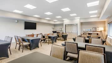 Un salón de conferencias de hotel con varias mesas y sillas, un televisor de pantalla plana y una zona de kitchenette.