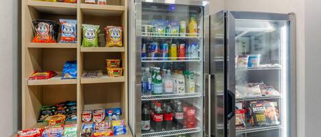 A kitchenette with a refrigerator stocked with beverages and snacks, and shelves with various food items.