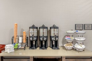 Una estación de bebidas con tres dispensadores de café, una pila de vasos naranja y un cartel que indica la ocupación máxima y la vestimenta adecuada.