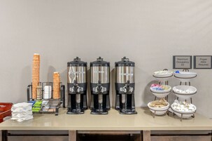 Una estación de bebidas con tres dispensadores de café, una pila de vasos naranja y un cartel que indica la ocupación máxima y la vestimenta adecuada.