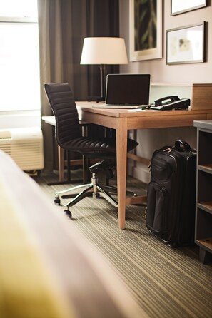 Habitación de hotel con un escritorio de madera, una silla de oficina negra, una computadora portátil, un teléfono y una maleta negra.