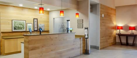 Un mostrador de recepción con detalles de madera, un quiosco digital de registro y una cámara de seguridad instalada en la pared.