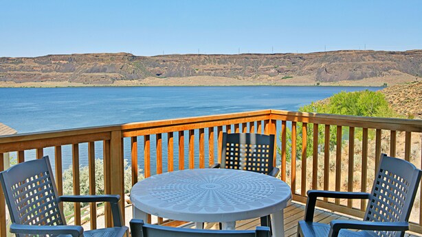 A deck with a table and chairs overlooking a body of water and a hilly landscape.