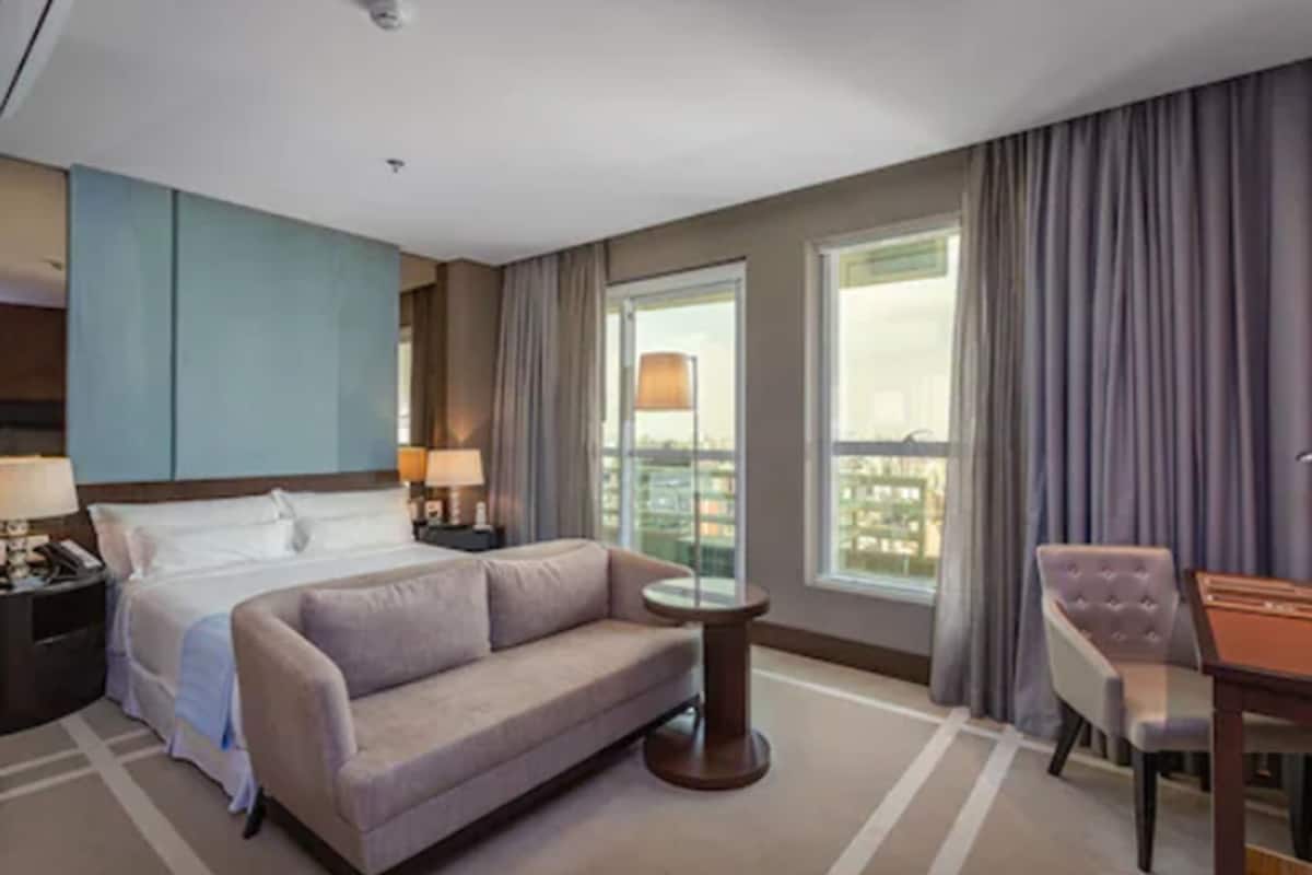 Quarto de hotel com cama, sofá, mesa pequena, cadeira e janela com cortinas.