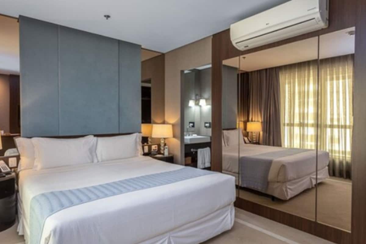 Quarto de hotel com uma cama grande, uma mesa de cabeceira com abajur, um espelho e um ar condicionado.