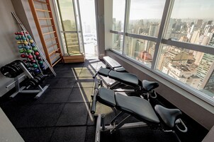 Una palestra con attrezzature per esercizi, una panca e vista sullo skyline urbano attraverso ampie vetrate.