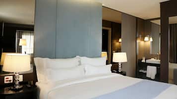 Habitación de hotel con una cama grande, dos lámparas de noche, una mesita de noche, un espejo, un baño con lavamanos y perchero, y una pared de color oscuro.